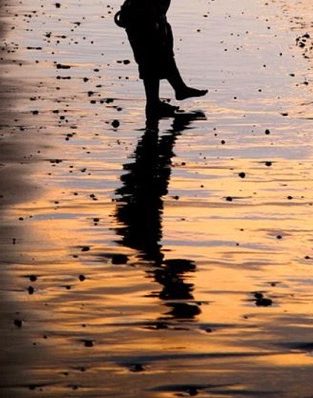 A person walking on a beach at sunset, their silhouette reflected in the wet sand.